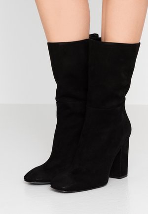 Schwarze Wildlederstiefel bis zur Mitte der Waden mit spitzen Zehen und klobigen hohen Absätzen, getragen von einer Person, die auf einem weißen Boden steht.