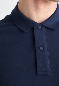 Mężczyzna w ciemnoniebieskiej koszulce polo z zapiętymi dwoma guzikami pod kołnierzykiem, kołnierzyk lekko rozłożony na boki.