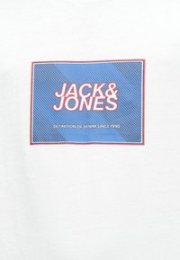 Biely tričko s modrou obdlžnikovou grafikou s diagonálnymi čiarami a červenými akcentmi, na ktorom je text "JACK & JONES" a "DEFINITION OF DENIM SINCE 1990."