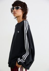 Osoba s kratkom kovrčavom kosom nosi bijele sportske naočale, crni oversized Adidas duksericu s bijelim trakama i burgundske hlače.