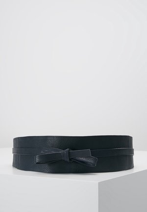 Brede zwarte leren riem met een smalle band die aan de voorkant in een strik is geknoopt, tentoongesteld op een wit voetstuk tegen een effen achtergrond.