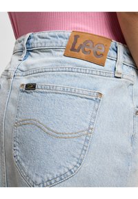 Des jeans en denim bleu clair présentent une patch en cuir marron avec la marque "Lee", des coutures visibles et un design classique à cinq poches.