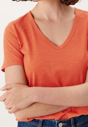 Kvinde iført en orange kortærmet V-hals shirt med armene krydsede, synlig fra halsen til taljen mod en ensfarvet baggrund.