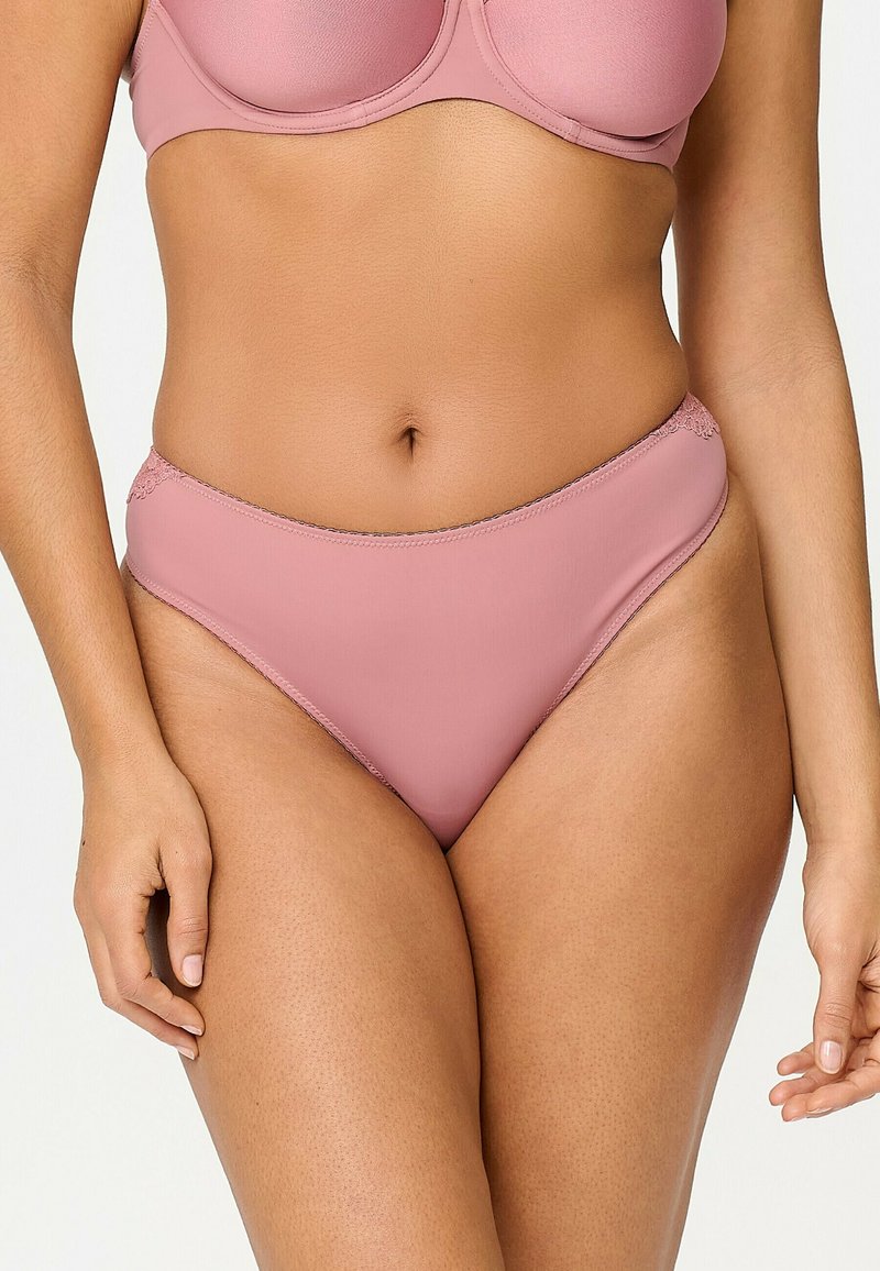 Femme portant un soutien-gorge rose pâle assorti et une culotte taille haute avec des détails en dentelle sur les hanches, visible de la moitié du torse jusqu'aux cuisses.
