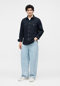 Homme portant une chemise noire à boutons, un jean large bleu clair et des baskets blanches, debout avec une main dans la poche.