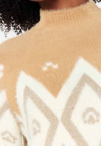 Pull en tricot duveteux présentant un motif géométrique en beige et crème avec des accents couleur fauve, col montant et texture douce.