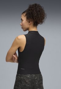 Γυναίκα με σγουρά μαλλιά σε ψηλή αλογοουρά φοράει μαύρη αμάνικη μπλούζα και σκουρόχρωμες γκρι κολάν με σχέδια, στέκεται με σταυρωμένα χέρια, προφίλ πλευράς.