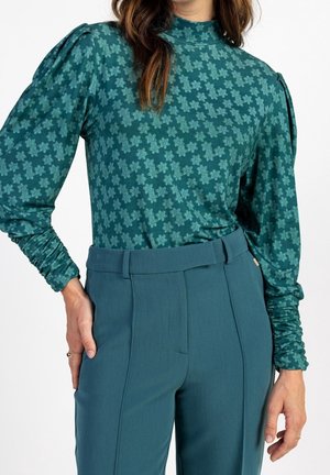 Frau in einer taillierten blauen Bluse mit floralen Mustern und Puffärmeln, die in hoch geschnittene, taillierte Hosen in Blau mit Gürtelschlaufen gesteckt ist.
