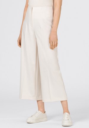 Henkilö pukeutunut leveälahkeisiin kermanvärisiin culottes-housuihin, ribbineulottuun hihattomaan toppiin ja valkoisiin lenkkareihin seisoo vaalean yksivärisen taustan edessä.