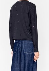 Top nero a maniche lunghe decorato con strass sparsi, abbinato a pantaloni in denim blu a gamba larga con tasche posteriori e cuciture visibili.
