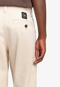 Beige byxor i en texturerad tyg, med en bakficka som har en svart logotyp patch och en brun knappdetalj.