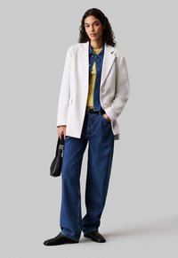 Biały oversize'owy żakiet, niebieska koszula dżinsowa, żółty sweter, szerokie niebieskie dżinsy, czarne skórzane buty oraz mała czarna torebka.