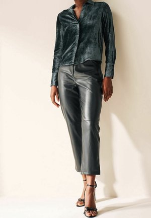 Personne portant une chemise texturée à manches longues foncée, un pantalon court en cuir noir et des sandales noires à talons hauts ouverts à l'avant, debout contre un mur clair.
