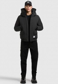 Juodas pūkų švarkas su kapišonu, su priekinėmis kišenėmis ir keturkampiu logotipo užsiuvimu. Nešiojamas su juodomis kroso kelnėmis ir juodais raištiniais batais.