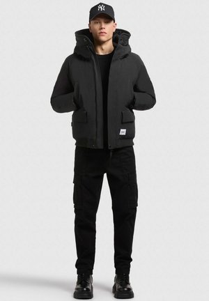 Zwarte pufferjack met een capuchon, met voorzakken en een vierkante logo-patch. Gekocht met zwarte cargobroeken en zwarte veterschoenen.