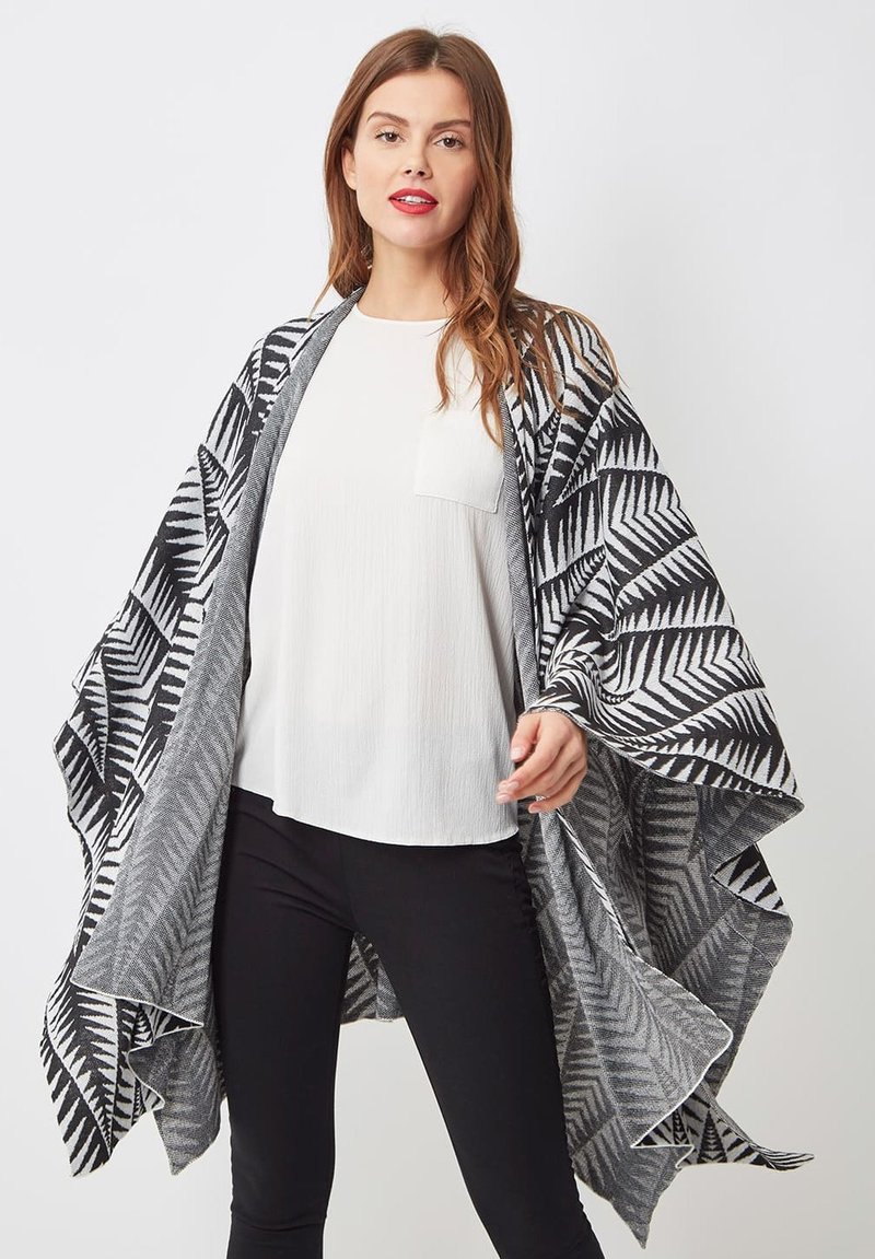 Mujer con un chal de patrón geométrico en blanco y negro sobre una blusa blanca y pantalones negros, de pie frente a un fondo liso.