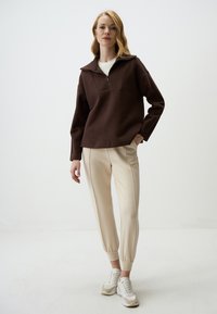 Felpa marrone con zip e ampio colletto, abbinata a pantaloni cream a gamba affusolata e sneakers bianche. Tessuto morbido con un design strutturato.