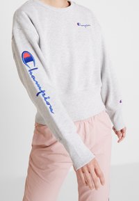 Sweat à capuche gris clair à manches longues. Présente le logo "Champion" brodé et des accents de design en rouge et bleu sur les manches.