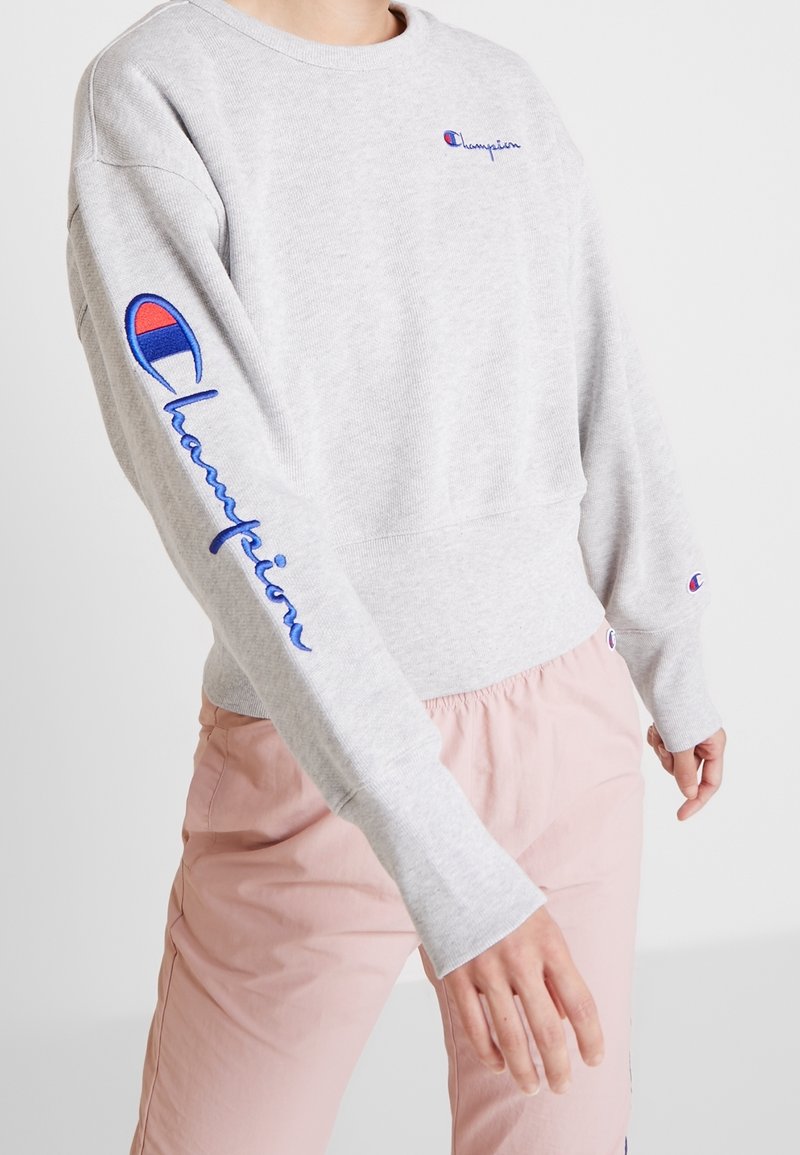 Sweat à capuche gris clair à manches longues. Présente le logo "Champion" brodé et des accents de design en rouge et bleu sur les manches.