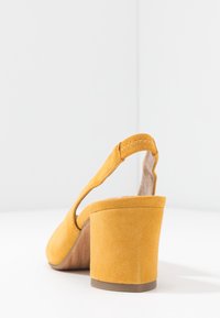 Tacco slingback in suede giallo con design curvo, superficie testurizzata e tacco spesso e robusto. Accentuato da dettagli di cucitura visibili.
