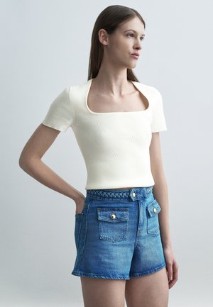 Kvinne iført en hvit ribbestrikket kortermet topp med firkantet hals og høytlivs jeansshorts med knapper foran på lommene, stående med den ene hånden i lommen.