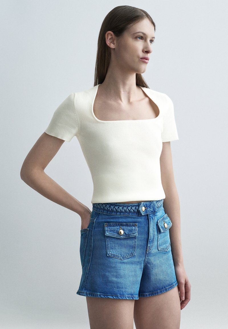 Γυναίκα που φοράει λευκό ριμπ μπλουζάκι με τετράγωνη λαιμόκοψη και κοντά μανίκια, καθώς και ψηλόμεσο τζιν σορτς με κουμπωμένες τσέπες στο μπροστινό μέρος, στέκεται με το ένα χέρι στην τσέπη.