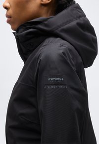 Personne portant une veste noire à capuche avec le logo Icepeak et le texte « It's out there » sur la partie supérieure de la manche, tourné vers la gauche sur un fond uni.