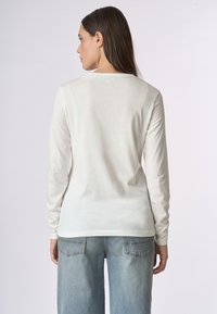Camicia bianca a maniche lunghe realizzata in materiale leggero con una texture liscia. Presenta un colletto girocollo semplice, indossata con jeans azzurri chiari.