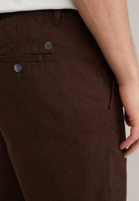 Los pantalones de algodón marrón cuentan con un bolsillo trasero con un detalle de botón negro y una textura sutil. No tienen patrones visibles ni acentos adicionales.