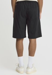 Sorte shorts lavet af let stof med elastisk taljebånd, der har en baglomme og en afslappet pasform, båret med hvide sokker og sko.