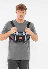 Svart och klar vattentålig bröstväska med justerbara remmar, med en framficka och ljusblå detaljer. Bärs av en person.