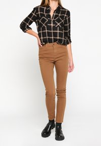 Žena stojící v krémových slim-fit kalhotách, černé a béžové kostkované košili s ohrnutými rukávy a černých robustních kotníkových botách na bílém pozadí.