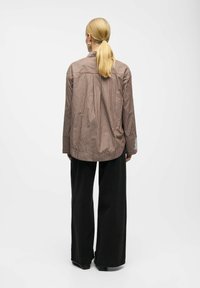 Brown and cream striped oversized shirt with a rounded hem, long sleeves, and white detailing at cuffs, paired with black wide-leg pants.