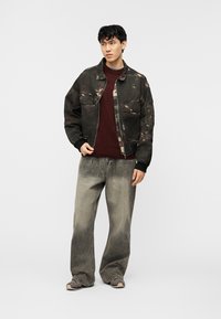 Νέος άνδρας με καμουφλαρισμένο στυλ μπουφάν μπόμπερ σε σκούρο χρώμα, βουργουνδί πουλόβερ, ξεθωριασμένο φαρδύ τζιν και γκρι αθλητικά παπούτσια, στέκεται μπροστά σε απλό φόντο.