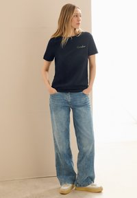 T-shirt en coton bleu marine à manches courtes et col rond ; présente un texte brodé vert. Associé à un jean large bleu clair et des baskets.