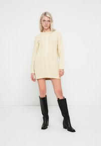 Φόρεμα από πλεκτό μάλλινο ύφασμα σε κρεμ απόχρωση με σχέδιο κοτσίδας, μακριά μανίκια και ριμπ τελείωμα. Φοριέται με μαύρες μπότες με τακούνι, που φτάνουν μέχρι το γόνατο.