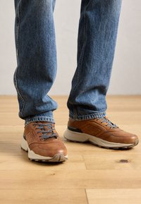 Personne portant un jean bleu et des baskets en cuir marron avec des lacets noirs et blancs, se tenant sur un sol en bois.