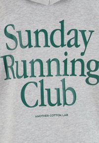 Felpa grigia con testo audace di colore verde scuro: "Sunday Running Club" sopra "ANOTHER COTTON LAB" in un carattere più piccolo. Morbido al tatto.