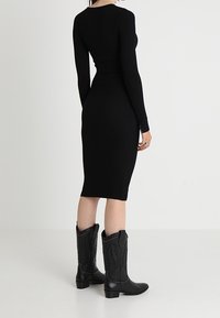 Abito nero a coste con maniche lunghe, silhouette aderente e orlo fino al ginocchio. Abbinato a stivali da cowboy neri con cuciture decorative.