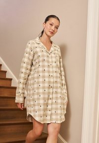 Béžový noční úbor s kostkovaným vzorem a malými zelenými motivy stromů, s knoflíkovým zapínáním vpředu, vykrojeným límcem a dlouhými rukávy.