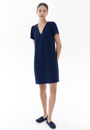 Marineblaues Kurzarmkleid mit V-Ausschnitt, aus glattem Stoff gefertigt. Verfügt über einen lockeren Schnitt und Seitentaschen, kombiniert mit dunklen Slippern.