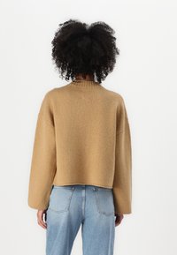 Maglione lavorato a maglia, corto, di colore cammello con una finitura texturizzata, dotato di un collo alto e maniche ampie. Abbinato a jeans blu chiaro.