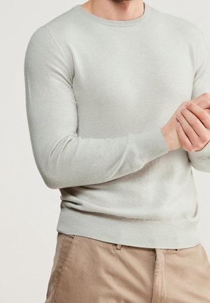 Homme portant un pull gris clair à manches longues et un pantalon beige, les mains jointes devant un fond uni.
