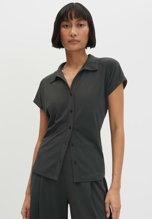 Femme aux cheveux noirs courts portant une chemise boutonnée à manches courtes de couleur sombre et un pantalon assorti, debout devant un fond clair uni.
