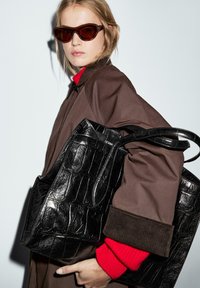 Borsa tote nera in pelle di coccodrillo impresso tenuta da una persona che indossa un cappotto oversized marrone e un maglione rosso con polsini a coste. Occhiali da sole con tonalità marrone.