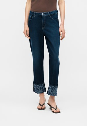 Woman wearing dark blue jeans with leopard print cuff detail and black wedge sandals, standing against a plain background.