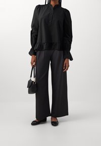 Donna che indossa una blusa nera larga con dettagli arricciati, pantaloni ampi scuri, ballerine nere e tiene una piccola borsa nera al suo fianco.