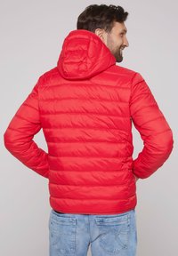 Rote Puffy-Jacke mit abnehmbarem Kapuzen, gestepptem Design und elastischen Bündchen. Über hellblauen Jeans getragen, die eine taillierte Passform zeigen.