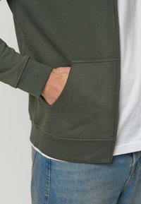 Hand in der Vorderhose einer grünen langärmligen Jacke, die über einem weißen Hemd und blauen Jeans getragen wird.