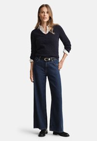 Marineblauer Strickpullover mit Kabelmuster, darunter ein weißes Hemd mit Kragen, kombiniert mit weitem, dunklem Denim-Jeans und schwarzen Ankle-Boots. Detail mit silberner Schnalle am Gürtel.